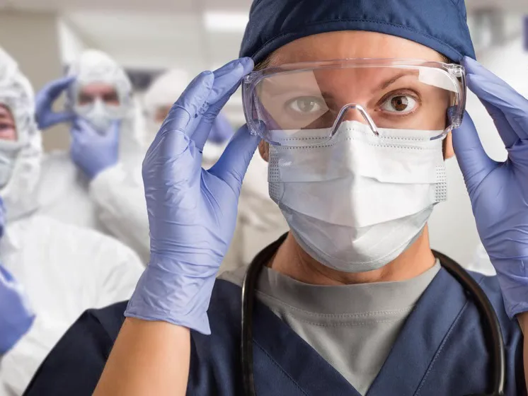 Healthcare worker adjusting safety goggles over a surgical mask, colleagues in protective suits behind